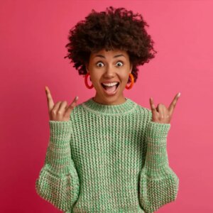 Happy carefree dark skinned rebellious young woman enjoys awesome music makes rock n roll gesture has fun on music festival or cool event wears casual jumper poses against pink wall